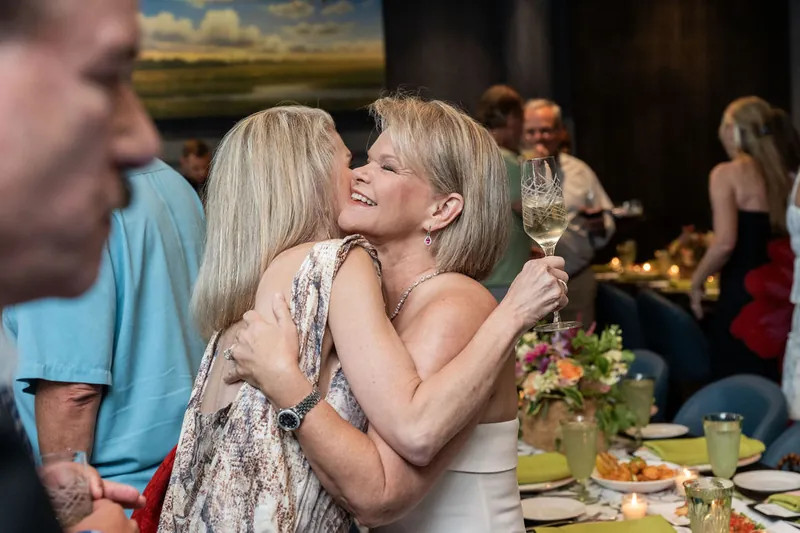 Two women embrace while holding wine glasses at a dining event, with guests and artwork visible behind them.