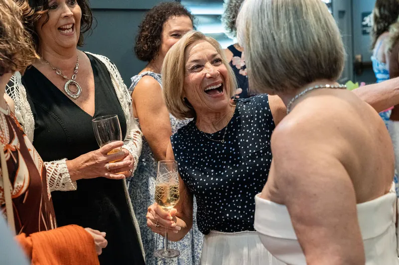 Three women laugh together during conversation at an indoor gathering, holding drinks and smiling warmly.