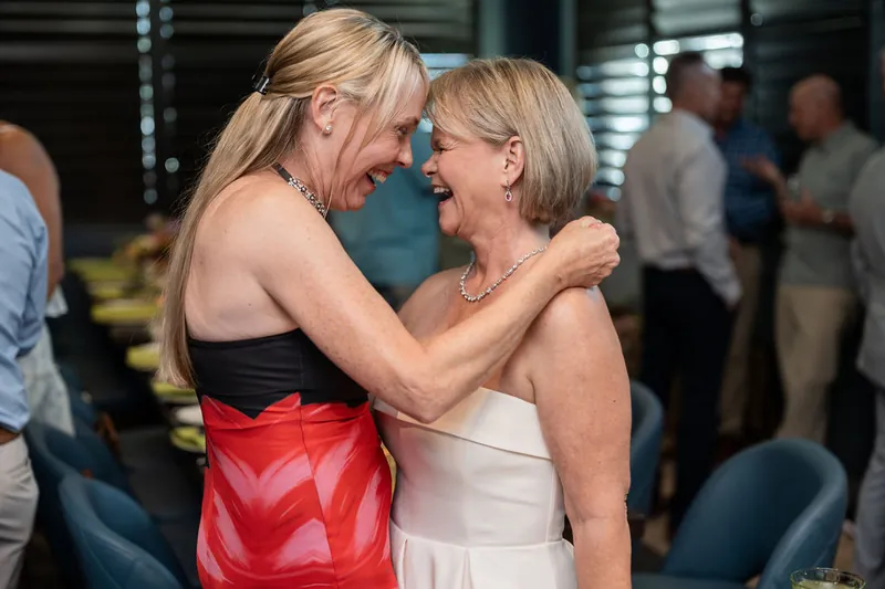Two women laugh while embracing at the reception, one in a red skirt and black top, the other in white.