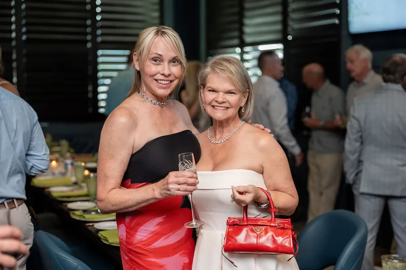 A woman in a black and red strapless dress holds a cocktail next to a woman in white holding a red clutch at the event.