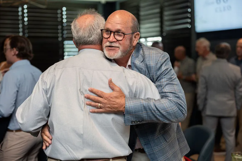 Two men embrace warmly during the celebration, one wearing a light blue shirt and the other in a darker button-up.
