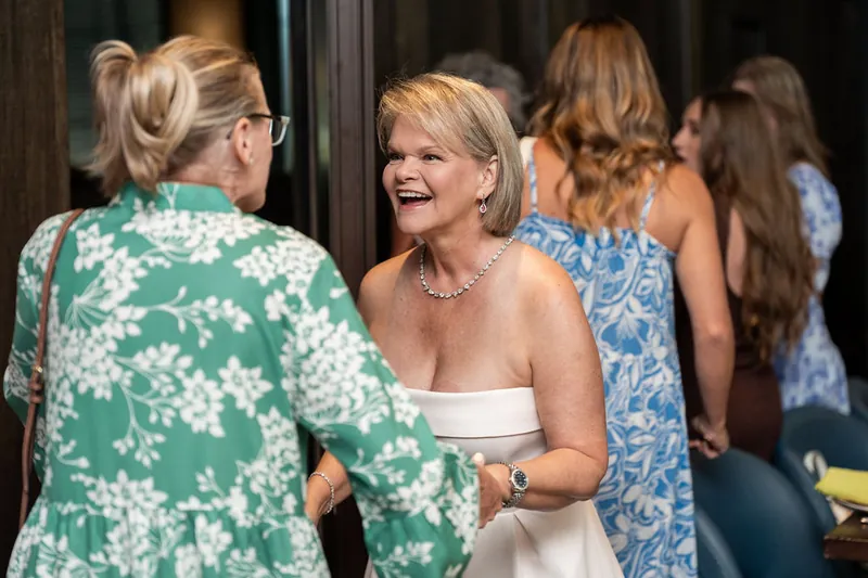 A woman in a white strapless dress smiles while chatting with guests in colorful floral prints at the indoor venue.