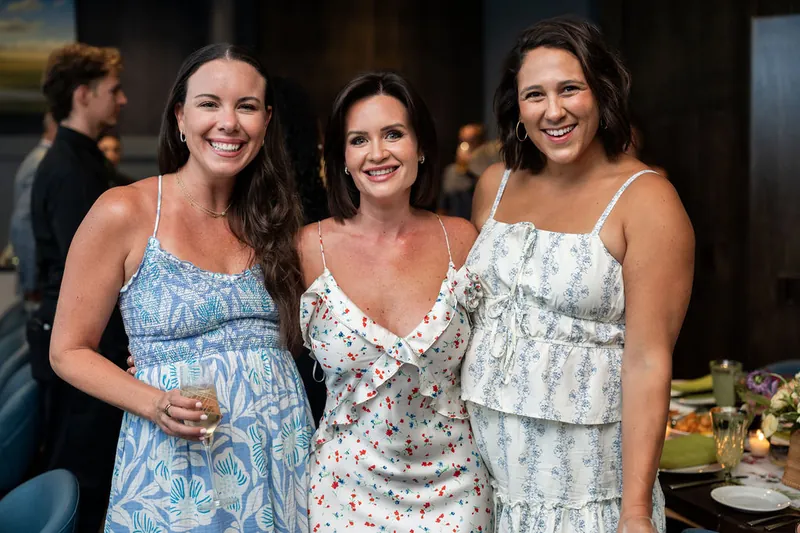Three young women stand together at the reception, each holding a drink and beaming at the camera in summery printed dresses.