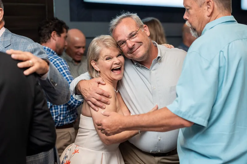 A man in a light blue shirt greets a woman in white at the event, surrounded by other guests.