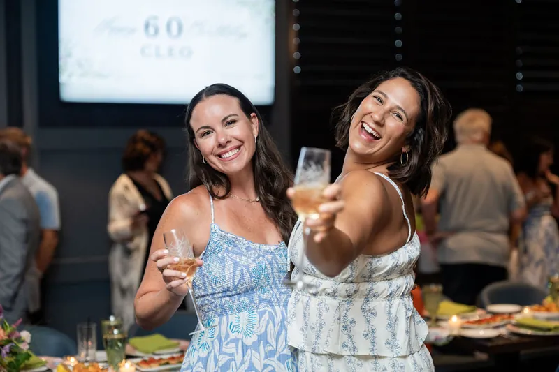 Two women laugh while holding drinks, with a projection screen visible behind them at the celebration.