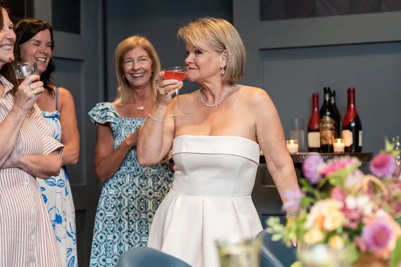A woman in a white strapless dress sips her drink while standing with friends, fresh flowers visible nearby.