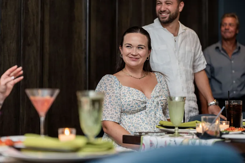 A woman in a floral dress sits at the table smiling as a man stands behind her during the gathering.