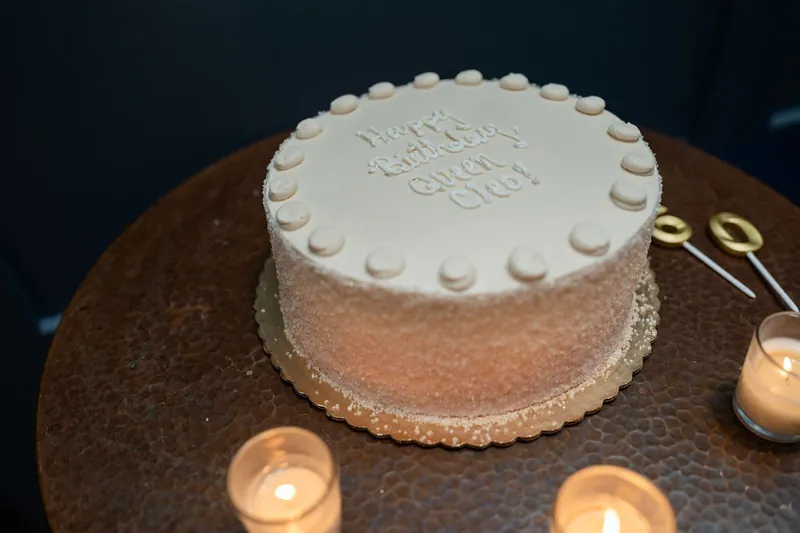 A white layer cake with piped border details sits on a dark table, "Happy Birthday Cleo" inscribed on top.