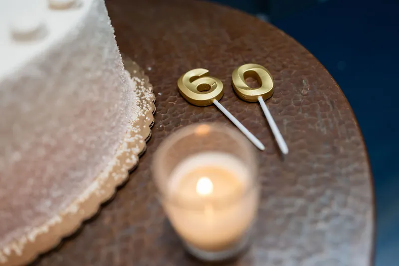 Gold number candle toppers rest on a dark surface beside a lit candle and tiered cake at the celebration.