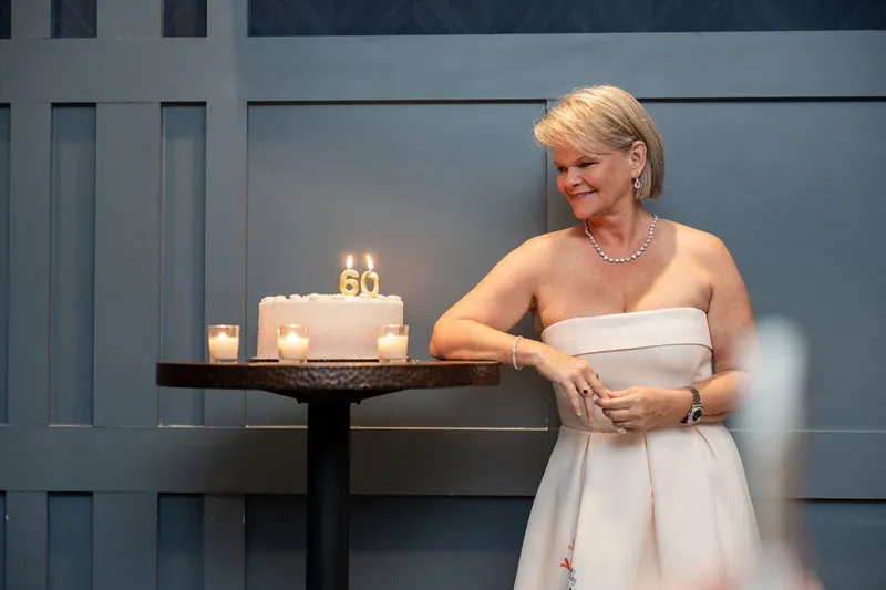 Cleo stands beside a black table holding a white tiered cake with gold accents and lit candles at Chophouse at Nocatee.