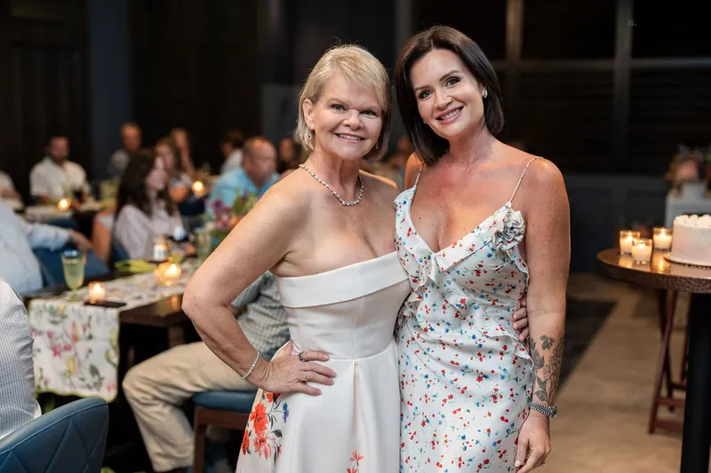 Two women smile together at the celebration, one in white and one in a floral dress.