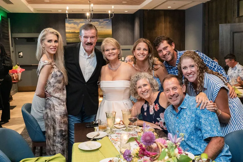 A group of nine family members poses together at their table, smiling at the camera with wine glasses and floral arrangements