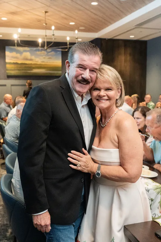 A man in a dark blazer and a woman in a white strapless dress embrace and smile for the camera during the celebration.
