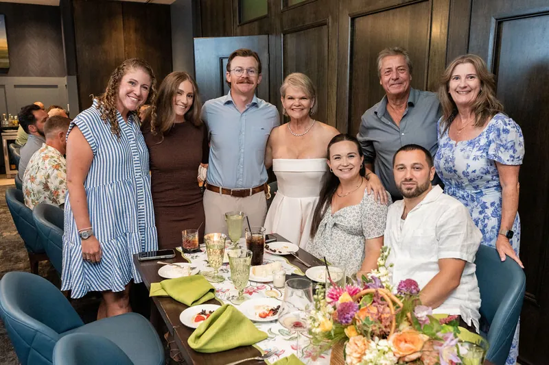 Eight relatives stand together at their dining table, with the woman in the center wearing a white sleeveless dress.