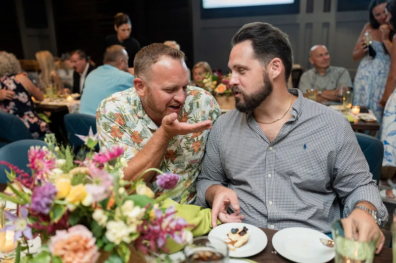 Two men sit at their table engaged in conversation, with vibrant pink and purple flowers and plated food in the foreground.