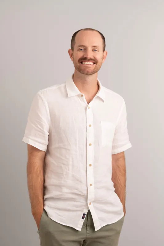 Man in white linen short-sleeve shirt smiling at camera against neutral gray backdrop.