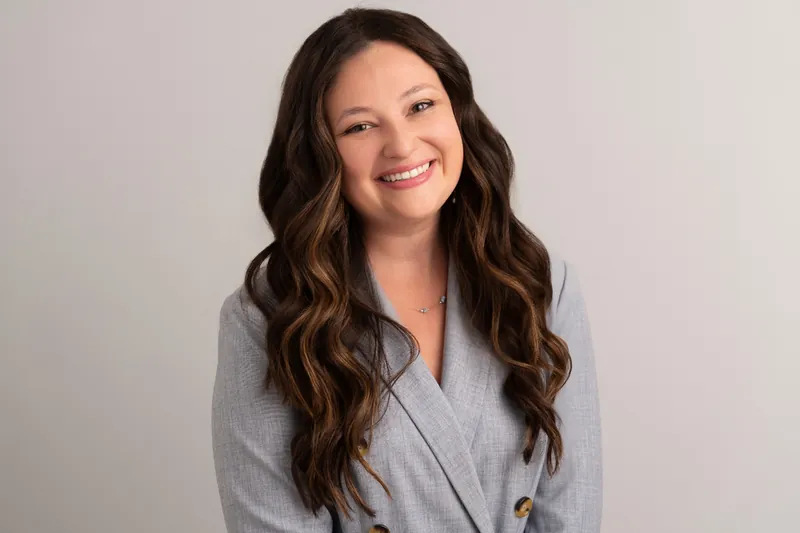 Woman with long dark wavy hair in gray blazer smiling warmly against neutral background.