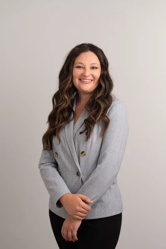 Woman in light gray cardigan with dark pants, hand on arm, smiling at camera.