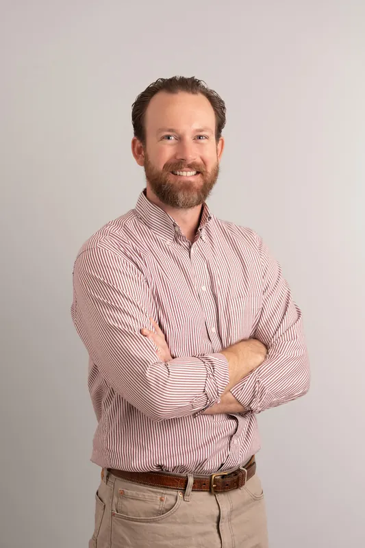 Man with reddish beard in pink striped shirt and khaki pants, arms crossed confidently.
