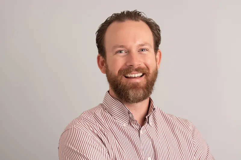 Bearded man in pink and white striped shirt grinning broadly against gray background.
