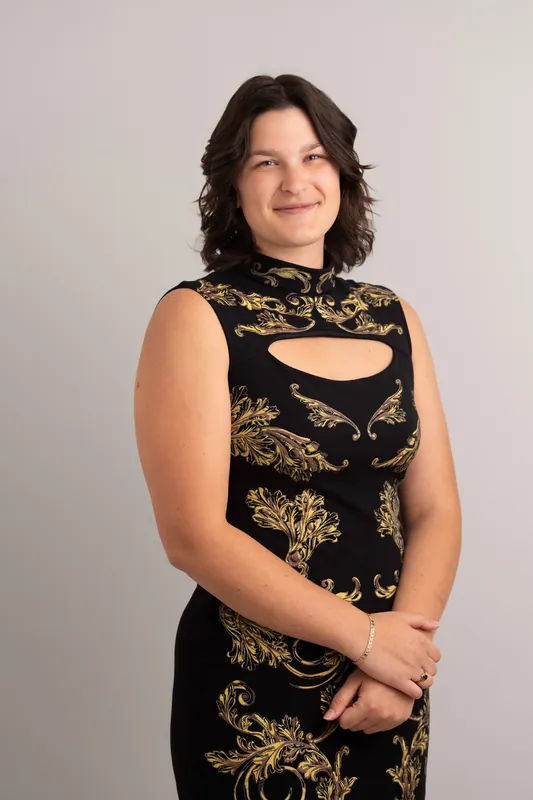 Woman with shoulder-length dark wavy hair wearing a black and gold patterned sleeveless top, hands clasped at her side.