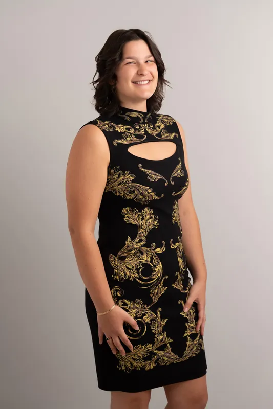 Woman in a black sleeveless dress with gold leaf pattern and cutout neckline stands against a neutral background