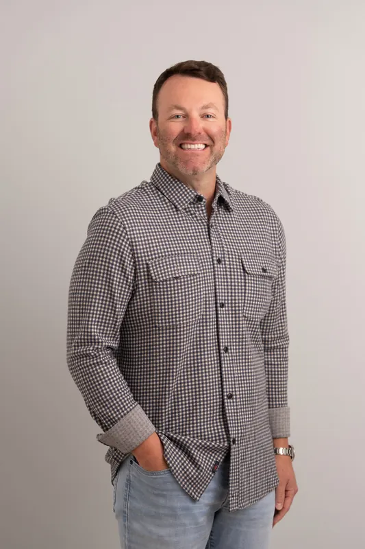 Tebault smiles at the camera in a gray gingham button-up shirt with one hand in his pocket against a white studio backdrop.