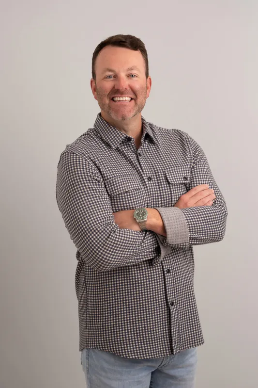 Tebault stands with arms crossed, wearing a gray checkered shirt and jeans, smiling confidently in the studio.