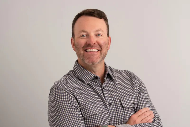 Tebault grins with arms crossed over his chest, dressed in a gray and white gingham shirt in the studio setting.
