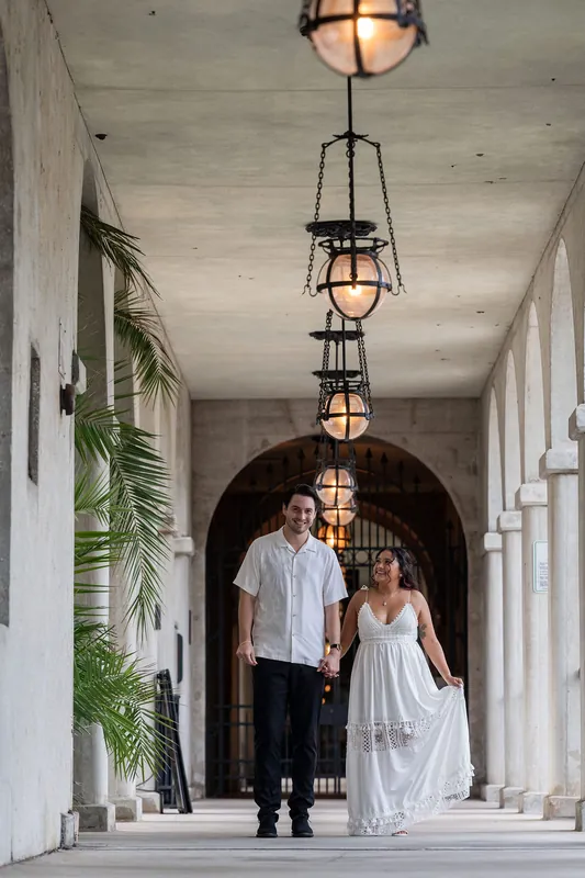 Dana and Christian walk hand in hand down a white-columned covered walkway with hanging lanterns and arched doorways.