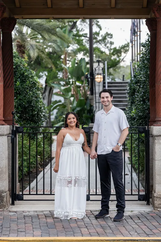 Dana and Christian stand at a wrought-iron gate framed by wooden pillars with lush greenery visible beyond the fence.