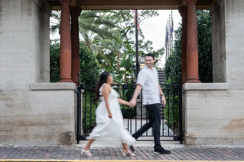 Dana in a white lace dress and Christian hold hands while walking through a covered passage with brick columns and iron