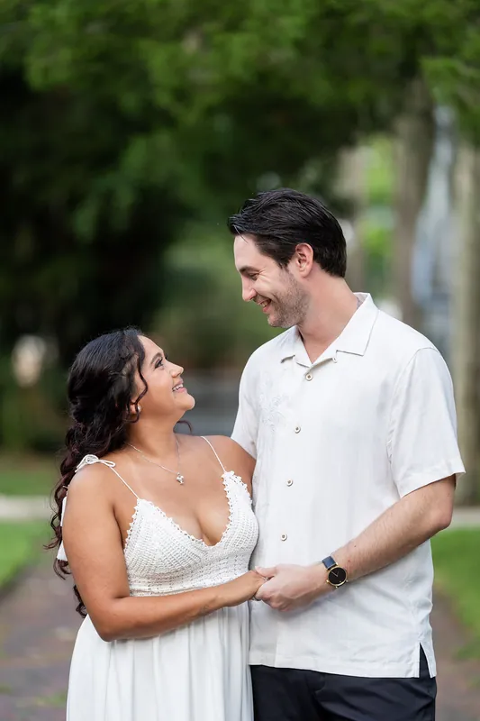 Dana in a white lace dress and Christian in a light button-up share a smile among green foliage along a tree-lined street.