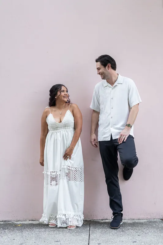 Christian and Dana stand against a pale wall; Dana wears a white eyelet dress while Christian holds his hat in dark pants.