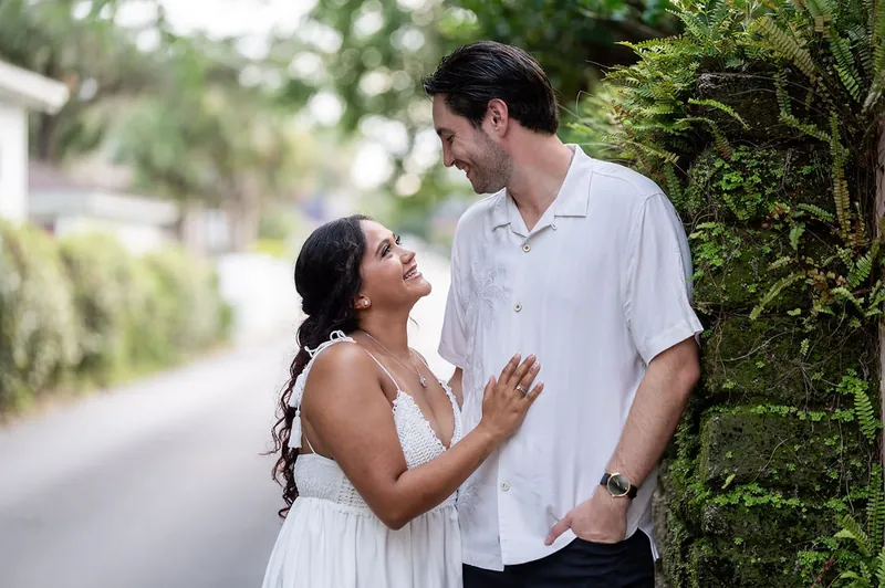 Dana gazes up at Christian as they walk together on a shaded residential path lined with hedges.