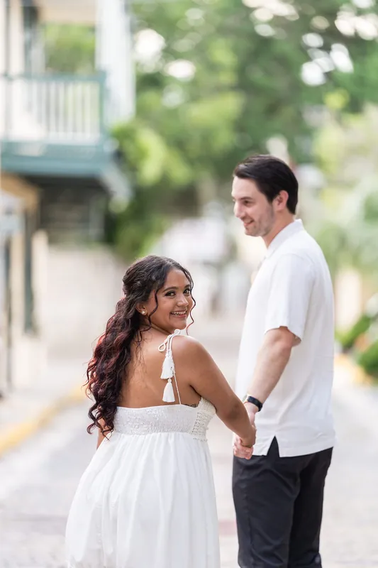 The couple holds hands on a quiet street with historic architecture visible behind them in soft afternoon light.