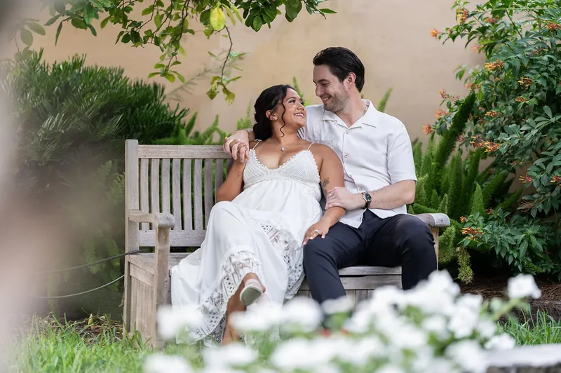 Dana and Christian sit close on a white bench surrounded by flowering plants and lush greenery.