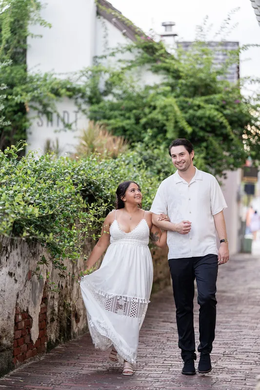 Walking arm-in-arm down a cobblestone passage, Dana's flowing white dress contrasts with ivy-covered stone walls.