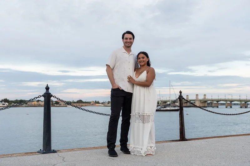 Dana and Christian pose together along the water's edge with boats and a bridge visible across the bay.