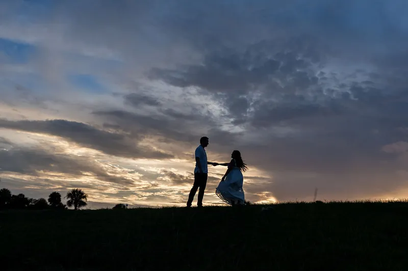 Silhouetted against a dramatic sunset sky, the couple stands close together in an open field.