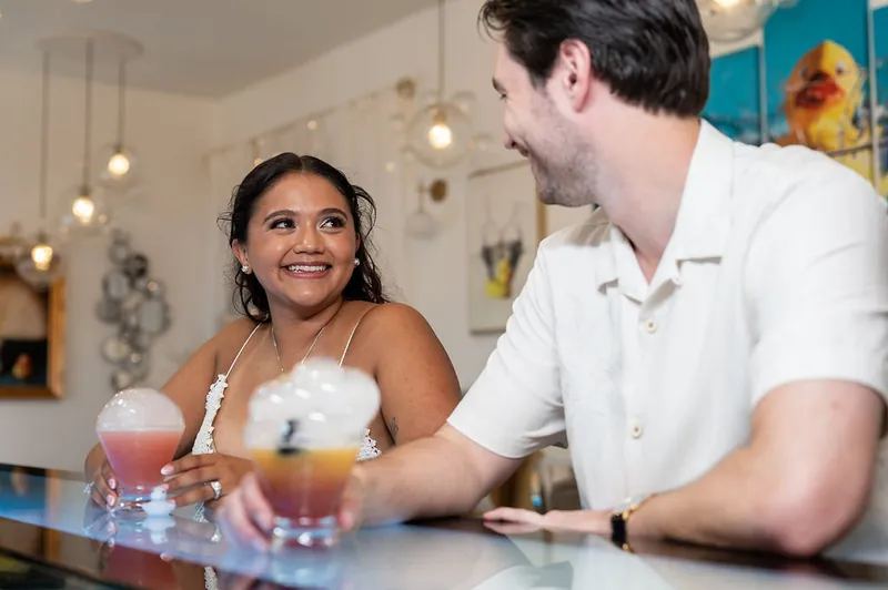 Dana smiles across the bar at Christian while holding a colorful cocktail at The Tipsy Duck Parlor.