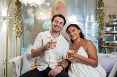 Dana and Christian clink glasses at The Tipsy Duck Parlor, both smiling as they sit together in front of holiday garland