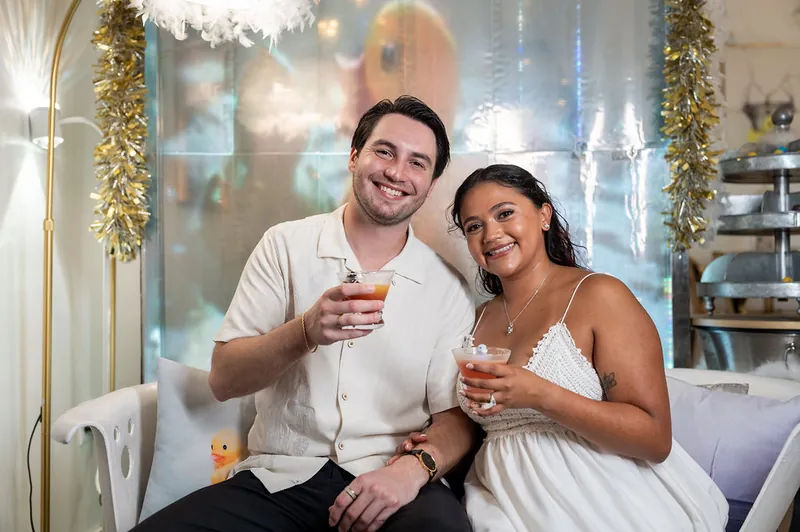 Dana and Christian clink glasses at The Tipsy Duck Parlor, both smiling as they sit together in front of holiday garland