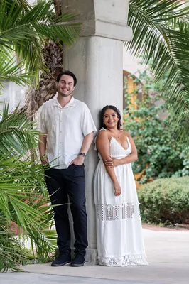 Christian and Dana stand together under a covered colonnade with palm fronds and tropical greenery surrounding them.