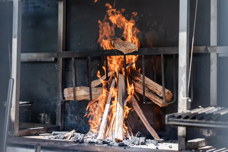 Blazing fire burns inside an outdoor Asado grill with stacked wood logs.