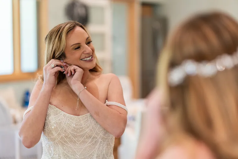 Danielle fastens pearl earring to her ear wearing beaded wedding dress.