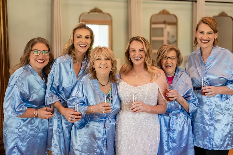 Danielle and five bridesmaids in matching blue patterned robes stand together smiling in a bridal suite with mirrors.