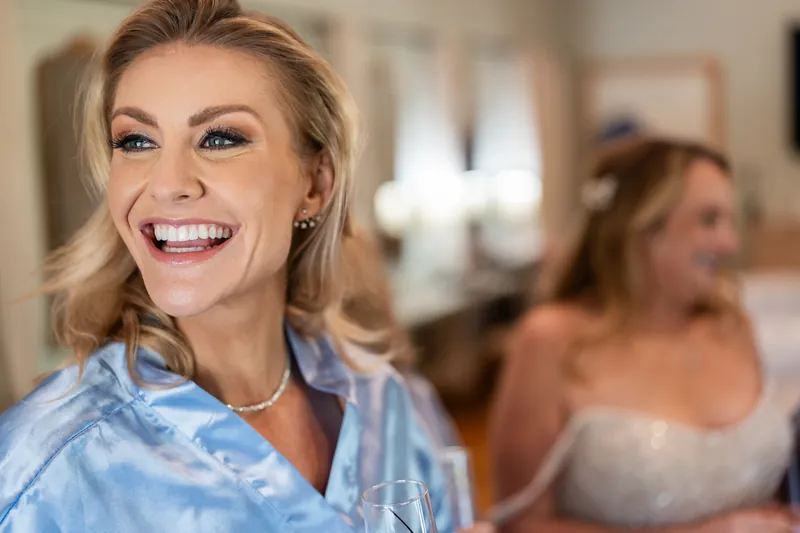 Close-up of a beaming bridesmaid in blue robe during pre-wedding festivities with guests blurred behind her.