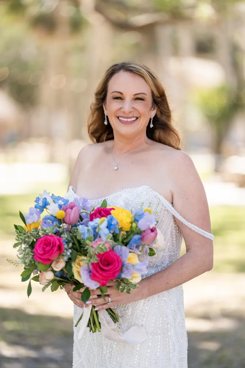Danielle smiles at the camera wearing a beaded white wedding dress and holding a colorful bouquet of blue, pink, and yellow flowers.