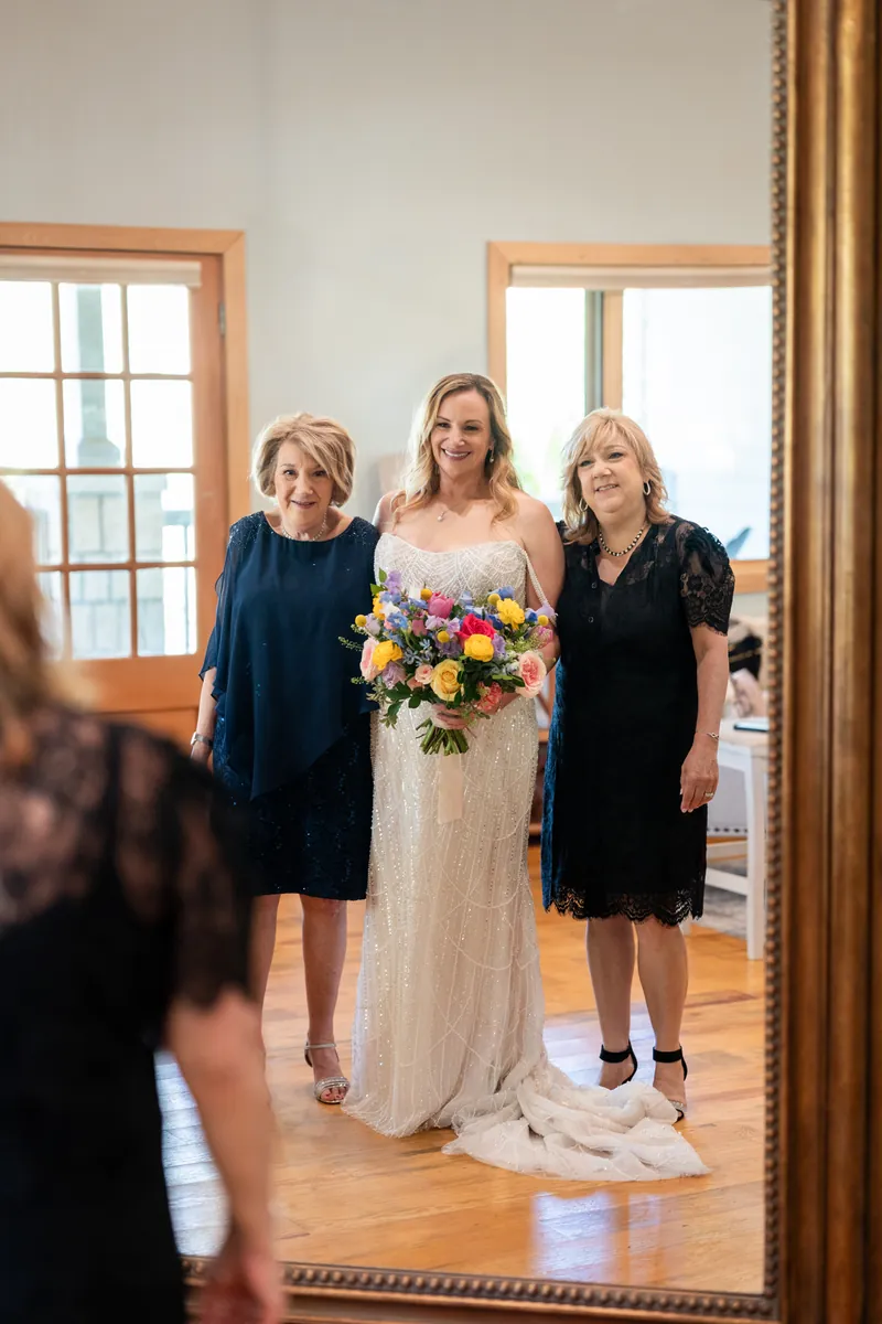 Danielle stands between two women in dark dresses, holding her vibrant mixed-flower bouquet indoors.